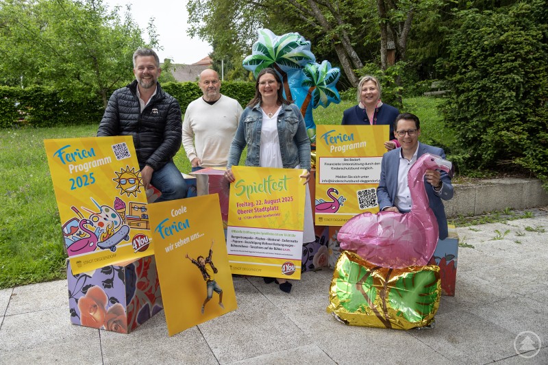 v.l.n.r.: Vorstellung des letztjährigen Ferienprogrammes – Sachgebietsleiter Bastian Löffelmann, Jugendbeauftragter Konrad Rankl, 4You Leiterin Karin Loibl, Vorsitzende des Kinderschutzbundes Ortsverband Deggendorf Plattling e. V. Yvonne Pletl-Schäfer und Oberbürgermeister Dr. Christian Moser Fünf Erwachsene stehen und sitzen im Freien vor einer Wiese und halten bunte Plakate für das Ferienprogramm 2025 und ein Spielfest. Sie sind von auffälligen Deko-Elementen wie einem Flamingo und einer aufblasbaren Palme umgeben.