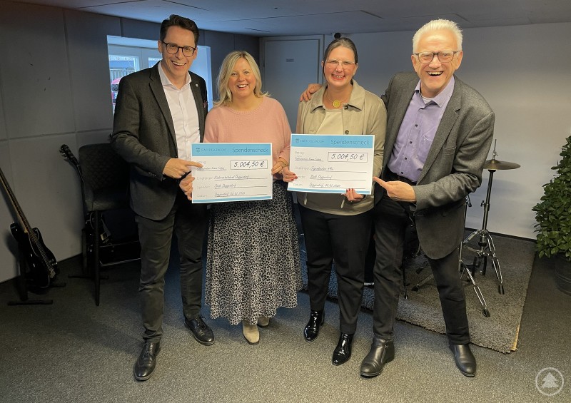 Oberbürgermeister Dr. Christian Moser, die Vorsitzende des Kinderschutzbundes Deggendorf Yvonne Pletl-Schäfer, die Leiterin des Jugendcenters 4You Karin Loibl und Generalmusikdirektor Johann Mösenbichler bei der Spendenübergabe aus dem Neujahrskonzert des Polizeiorchesters Bayern. Vier Personen stehen nebeneinander in einem Innenraum und halten zwei große Spendenschecks der Stadt Deggendorf in den Händen. Alle Beteiligten lächeln in die Kamera. Im Hintergrund sind Musikinstrumente zu sehen, was auf einen kulturellen Rahmen der Spendenübergabe hinweist.