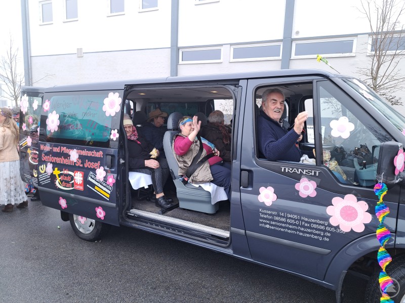 Mit dem bunt dekorierten Kleinbus beteiligte sich das Seniorenheim St. Josef am Faschingszug in Hauzenberg. in geschmückter Kleinbus des Seniorenheims St. Josef mit Hippie Blumen fährt beim Faschingszug durch Hauzenberg, mehrere Seniorinnen und Senioren winken fröhlich aus dem Fahrzeug.