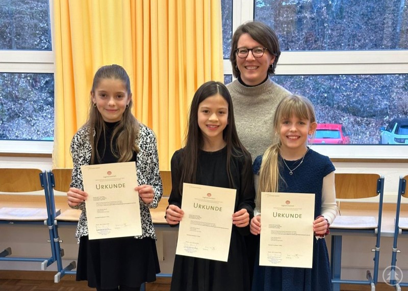 Das junge Querflötentrio Anna Stadler (vorne, v. l.), Amalie Hartl und Antonia Burkert wird betreut von Lehrerin Elisabeth Wagner-Thoma (hinten). Zwei Schülerinnen halten ihre Urkunden in die Kamera, hinter ihnen steht ihre Lehrerin in einem hellen Musikschulraum mit großen Fenstern und gelben Vorhängen.