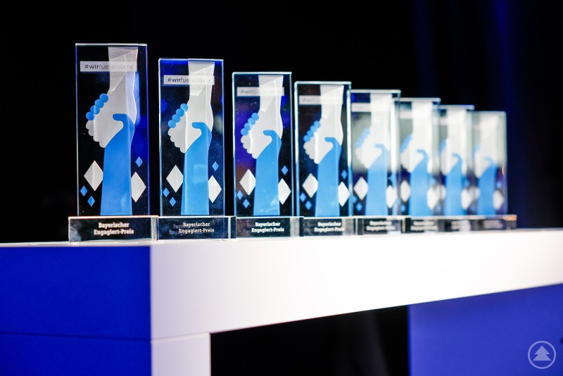 Bayerischer Engagiert-Preis. Mehrere gläserne Auszeichnungen stehen in einer Reihe auf einem Tisch, im Hintergrund ist eine Bühne mit blauem Licht zu sehen.