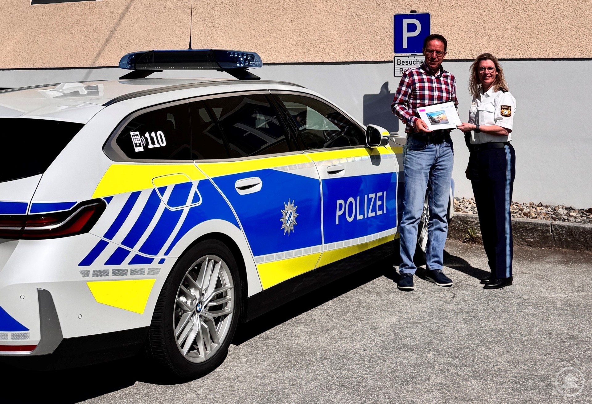 PORin Ingrid Grötzinger übergibt Bürgermeister Gerhard Poschinger vor dem Rathaus von Perlesreut den aktuellen Sicherheitsbericht. Vor dem Rathaus von Perlesreut stehen ein Polizeiauto und zwei Personen – eine Polizeibeamtin in Uniform und ein Mann mit Hemd und Jeans. Sie halten gemeinsam ein Dokument in der Hand und lächeln in die Kamera.