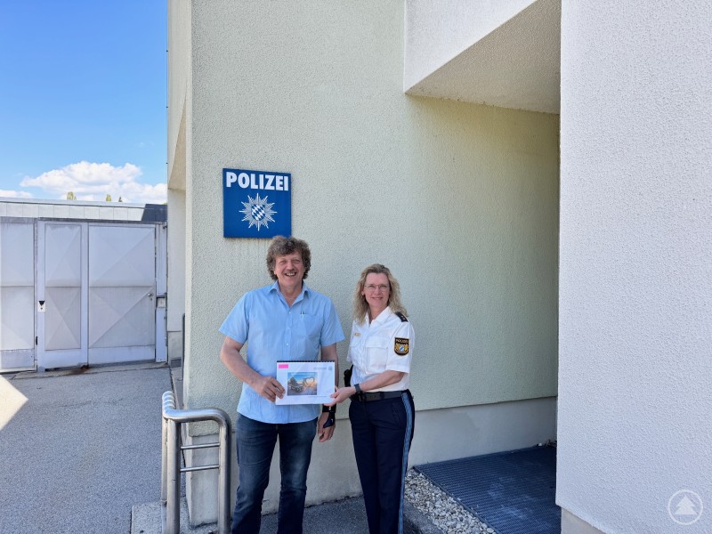 PORin Ingrid Grötzinger übergibt Bürgermeister Helmut Knaus den Sicherheitsbericht für die Gemeinde Philippsreut. Ein Mann im blauen Hemd und eine Polizeibeamtin in Uniform stehen lachend vor dem Polizeigebäude unter einem Schild mit der Aufschrift „Polizei“. Beide halten ein Dokument mit Titelbild in der Hand.