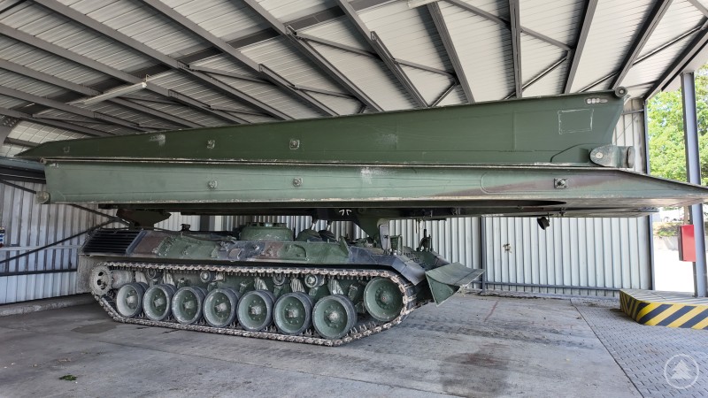 Ein grüner Brückenlegepanzer steht unter einem Blechdach. Die aufgelegte Brückenkonstruktion ist eingefahren, das Kettenfahrzeug darunter steht einsatzbereit.