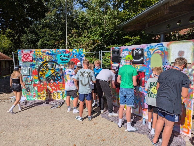 Mehrere Jugendliche gestalten bunte Graffitis auf einer großen Wand im Freien. Sie sprühen farbenfrohe Motive und Schriftzüge, während die Sonne scheint.