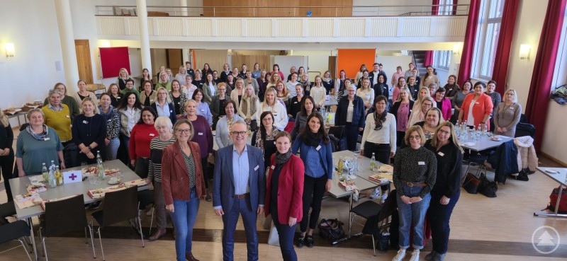 Die Caritas-Kitas sind eine starke Kraft in Kirche und Gesellschaft. Die Leiterinnen der über 100 Einrichtungen mit den Caritasvorständen, Direktorin Andrea Anderlik, Bischöflich Beauftragter Stefan Seiderer und der Kita-Abteilungsleiterin Melanie Krieger im Vordergrund. Eine große Gruppe von etwa hundert Personen steht in einem hellen Saal mit hohen Fenstern und roten Vorhängen. Im Vordergrund posieren mehrere Personen für das Gruppenfoto, während im Hintergrund Tische mit Getränken und Unterlagen zu sehen sind.