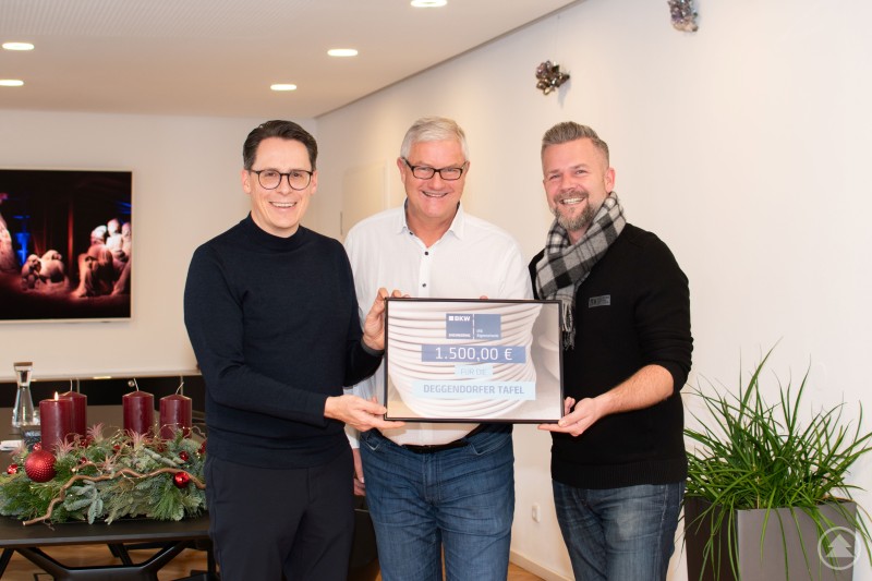 Oberbürgermeister Dr. Christian Moser, Dipl.-Geol. Dr. rer. nat. Roland Kunz (Geschäftsführer von IFB Eigenschenk) und Bastian Löffelmann (Sachgebietsleiter Schulen, Kinderbetreuung, Soziales und Sport) bei der Spendenübergabe. Drei Männer stehen lächelnd in einem modern eingerichteten Raum und halten gemeinsam ein großes Spendenschild über 1.500 Euro für die Deggendorfer Tafel. Im Vordergrund steht ein festlicher Adventskranz mit Kerzen, im Hintergrund sind ein Bild und Pflanzen zu sehen.