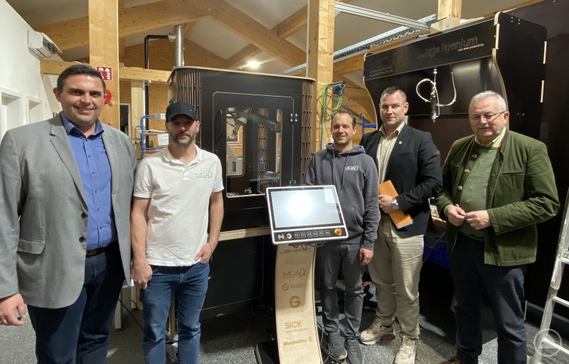 Bürgermeister Andreas Urban, Matthias und Christoph Wolf, Wirtschaftsförderer Martin Köck und Landrat Josef Laumer bei einem Rundgang durch die Produktionsräume der MCW Systemtechnik GmbH in Wiesenfelden. Mehrere Personen stehen in einer Produktionshalle vor einer modernen Roboterzelle und einer Steuerkonsole. Im Hintergrund sind Holzbalken, Maschinen und technische Anlagen zu sehen. Die Gruppe posiert für ein Foto während eines Firmenbesuchs.