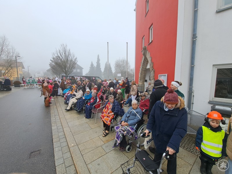 Zahlreiche Seniorinnen und Senioren sitzen eingeh&uuml;llt in Decken vor dem Seniorenheim am Stra&szlig;enrand und verfolgen bei nebligem Wetter den Faschingszug in Hauzenberg.