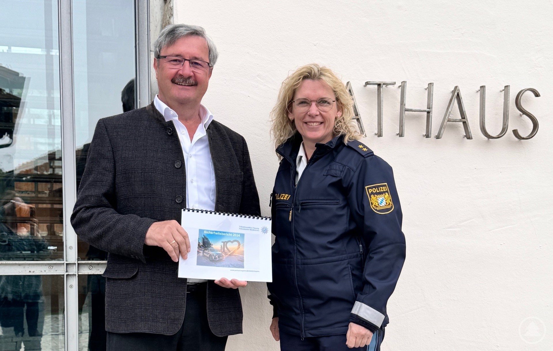 PORin Ingrid Grötzinger überreicht Bürgermeister Fritz Raab vor dem Rathaus Hinterschmiding den Sicherheitsbericht. Ein Mann in dunkler Jacke und eine Polizeibeamtin in Uniform stehen nebeneinander vor dem Rathaus. Beide halten gemeinsam ein Dokument und lächeln in die Kamera.
