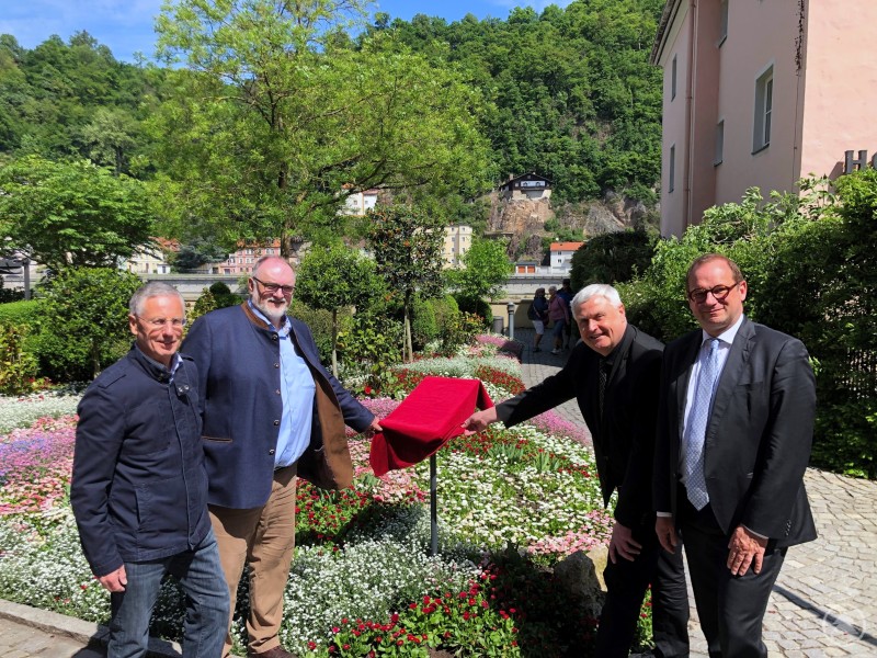 Oberbürgermeister Dupper (2. v. l.) enthüllt zusammen mit dem Ideengeber Prof. Dr. Harnisch (li.), Dompropst Dr. Bär (2. v. r.) und dem Kulturreferenten der Stadt Passau, Dr. Forster (re.), das Schild am Tencalla-Garten. Vier Männer stehen in einem blühenden Garten, drei von ihnen heben ein rotes Tuch an einem Schild. Im Hintergrund sind Altstadthäuser und eine bewaldete Anhöhe zu sehen.