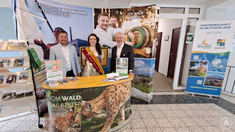 Erster Bürgermeister der Glasstadt Zwiesel Karl-Heinz Eppinger, Glaskönigin Susanne Glanzner und Robert Kürzinger, Geschäftsführer der Ferienregion Nationalpark Bayerischer Wald, präsentieren die Region beim Rathausfest in Brake. Drei Personen stehen an einem bunt gestalteten Messestand mit Prospektmaterialien vor zwei großen Bannern der Ferienregion Nationalpark Bayerischer Wald. Eine Frau trägt eine Tracht und eine Schärpe, zwei Männer stehen rechts und links von ihr. Im Hintergrund ist ein Roll-Up mit der Städtepartnerschaft Brake – Zwiesel zu sehen.