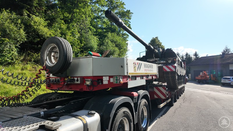 Eine Panzerhaubitze 2000 mit langem Geschützrohr wird auf einem Tieflader der Firma Wagner transportiert. Die Szene spielt sich an einer Straße mit Wald im Hintergrund ab.