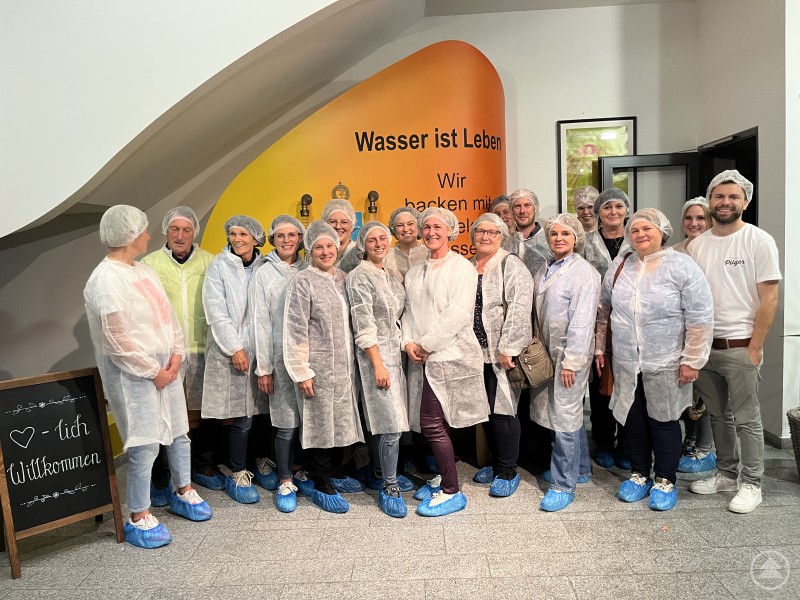 Die Teilnehmenden erhielten bei einer ausführlichen Führung durch Inhaber David Pilger (rechts) spannende Einblicke in die Abläufe einer modernen Handwerksbäckerei. Eine Gruppe von rund 20 Personen steht in Schutzkleidung mit Haarnetzen und blauen Überziehschuhen in einer Bäckerei. Im Hintergrund ist eine gelbe Wand mit der Aufschrift „Wasser ist Leben – Wir backen mit reinstem Bergquellwasser“ zu sehen. Alle lächeln in die Kamera.