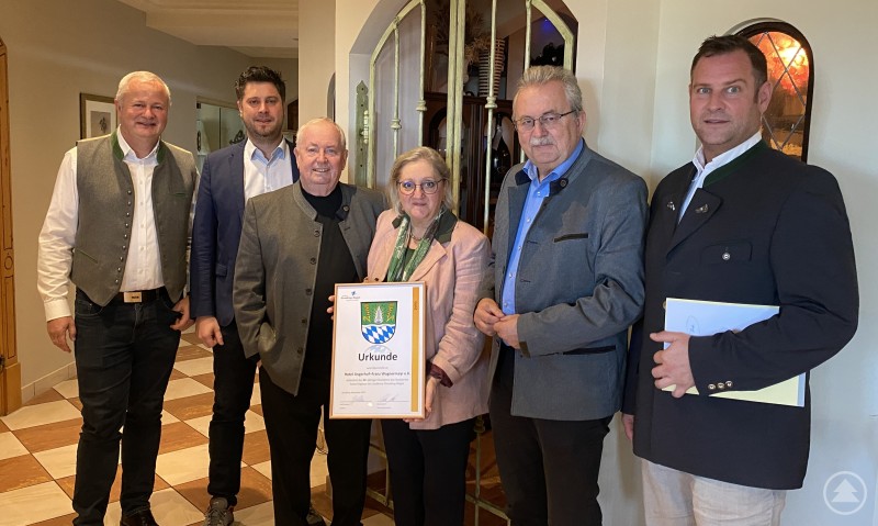 Landrat Josef Laumer gratulierte Franz und Maria Wagnermayr zum 40-jährigen Bestehen des Hotels Angerhof in Sankt Englmar. Sechs Personen stehen in einem Gastraum zusammen und halten eine gerahmte Urkunde. Im Hintergrund ist eine verzierte Türöffnung zu sehen.