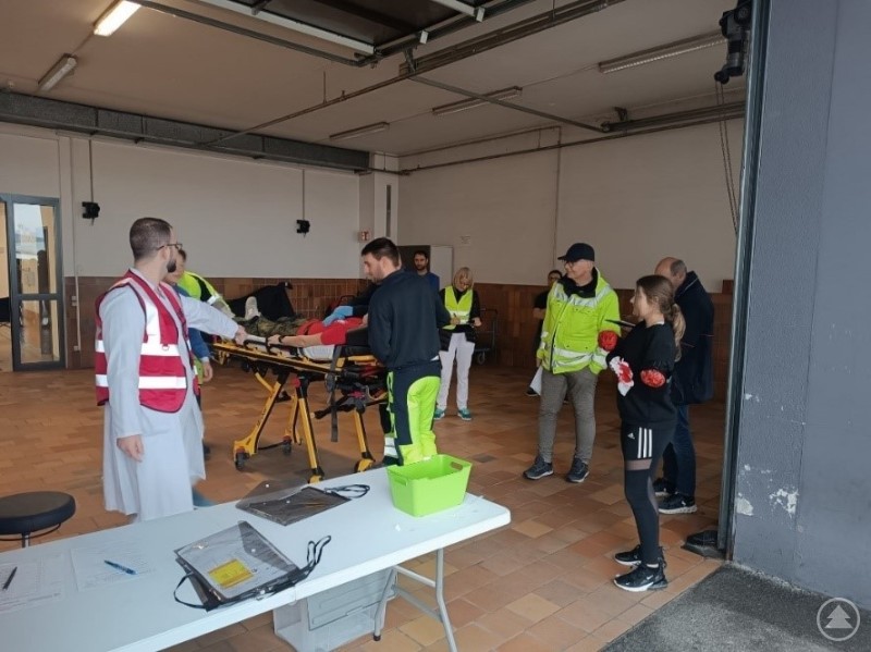 Einsatzkräfte und Klinikmitarbeiter üben die Triage und Erstversorgung bei einem angenommenen Massenanfall von Verletzten. Mehrere Einsatzkräfte und Übungsteilnehmende stehen um eine Person auf einer Trage in einer Garage oder Halle, während ein Tisch mit Unterlagen im Vordergrund steht.