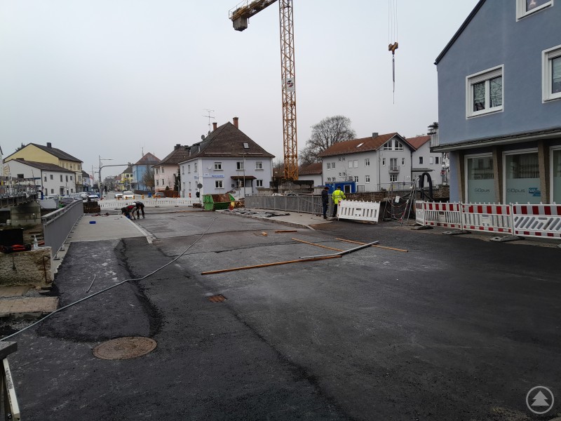Die neue Bogenbachbrücke in Bogen wird am 19. Dezember wieder für den Verkehr freigegeben. Eine frisch asphaltierte Brücke mit Absperrgittern und Bauarbeiten im Stadtgebiet von Bogen. Im Hintergrund sind Baukran, Bauarbeiter und angrenzende Wohnhäuser zu sehen. Die Straße ist für den Verkehr noch gesperrt.