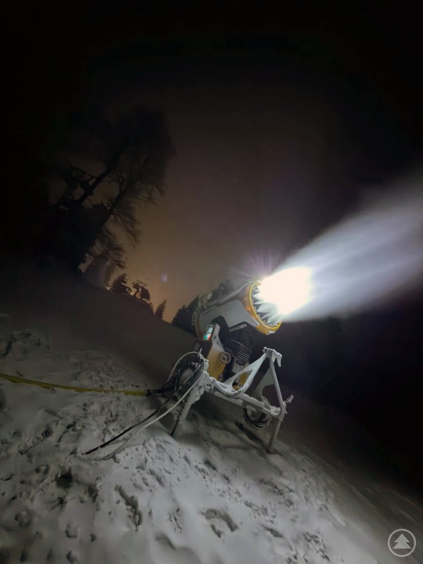 Eine Schneekanone steht nachts auf einer mit frischem Schnee bedeckten Piste und spr&uuml;ht intensiv Schnee in die Dunkelheit. Im Hintergrund sind B&auml;ume und schwache Lichtquellen zu erkennen.