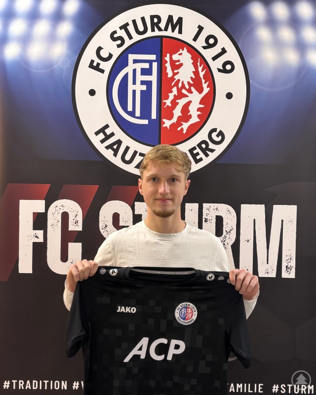 FC Sturm Hauzenberg verpflichtet Leandro Miller. Ein junger Fußballspieler steht vor dem Vereinslogo des FC Sturm Hauzenberg und hält ein schwarzes Trikot mit Sponsorenaufdruck in die Kamera.