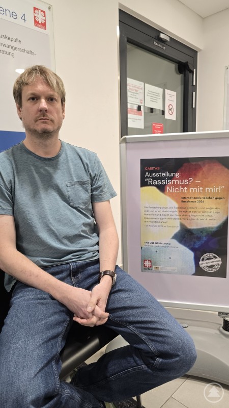 Andreas Trpak hat die Ausstellung „Rassismus? Nicht mit mir.“ konzipiert und realisiert. Sie ist im Treppenhaus des Konradinums in Passau zu sehen und begleitet die Internationalen Wochen gegen Rassismus. Ein Mann sitzt neben einer Ausstellungstafel der Caritas mit dem Titel Rassismus Nicht mit mir. Die Tafel steht im Treppenhaus des Konradinums in Passau und informiert über die Internationalen Wochen gegen Rassismus.