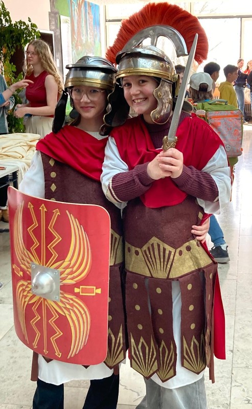 In römischen Kostümen wurde die Antike beim Römertag am LLG lebendig. Zwei Schülerinnen tragen römische Kostüme mit Helmen, roten Umhängen, Schild und Schwert und posieren in der Aula des Gymnasiums für ein Foto.