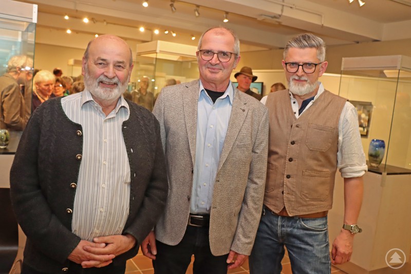 Freuen sich über eine gelungene Ausstellung: Laudator Ossi Heindl (von links), Künstler Rainer Metzger und Museumsleiter Christian Binder. Das Bild zeigt drei Männer, die in einem Ausstellungsraum oder Museum stehen. Sie tragen größtenteils traditionelle oder schlichte, elegante Kleidung – darunter ein Janker, ein Sakko und eine Weste. Im Hintergrund sind Vitrinen mit Ausstellungsstücken sowie weitere Personen zu sehen, was auf eine Veranstaltung oder Ausstellungseröffnung hindeutet. Die Atmosphäre wirkt freundlich und interessiert.