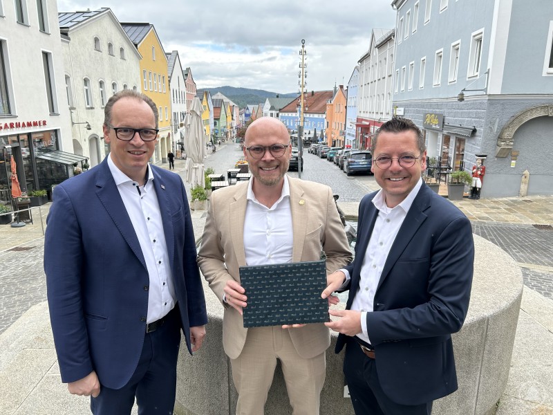 Gastgeschenke empfehlen sich weiterhin eher analog. Bürgermeister Heinz Pollak (v.r.) freute sich, Digitalminister Fabian Mehring und MdL Martin Behringer in Waldkirchen willkommen heißen zu dürfen. Drei Männer im Anzug stehen lächelnd vor einem Brunnen in der Altstadt von Waldkirchen, im Hintergrund sind bunte Häuserfassaden und ein Maibaum zu sehen.