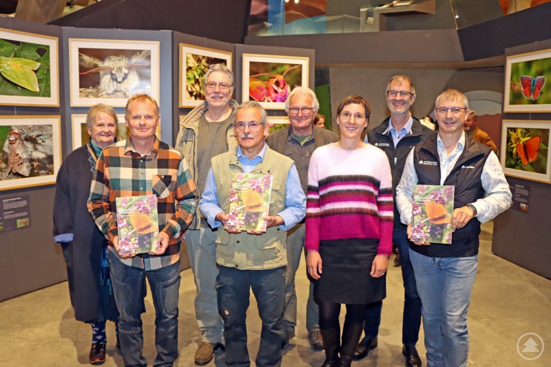 Die Macher des Schmetterlingsbands mit Nationalparkleiterin Ursula Schuster (3.v.r.) inmitten der beeindruckenden Fotos der mittlerweile zu Ende gegangenen Ausstellung im Haus zur Wildnis. Eine Gruppe von acht Personen steht lächelnd vor einer Ausstellung mit großformatigen Schmetterlingsfotos. Einige halten ein Buch in den Händen. Im Hintergrund sind gerahmte Natur- und Insektenaufnahmen an den Wänden zu sehen.
