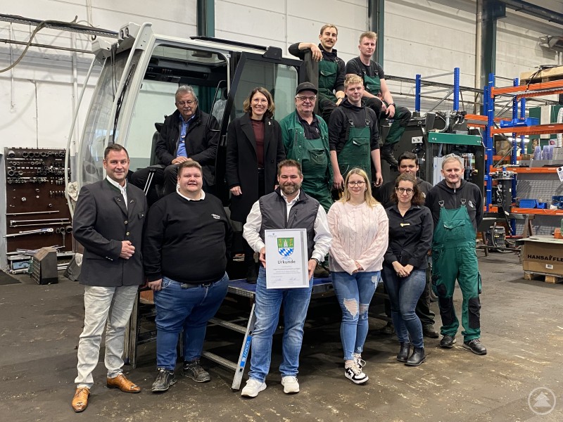 Landrat Josef Laumer besuchte gemeinsam mit Bürgermeisterin Andrea Probst die Impex Forstmaschinen GmbH in Bogen anlässlich des 25-jährigen Jubiläums. Eine Gruppe von Mitarbeitenden und Gästen steht in einer Werkhalle vor einer großen Forstmaschine. Eine Person hält eine Urkunde, im Hintergrund sind Werkzeuge und Regale zu sehen.