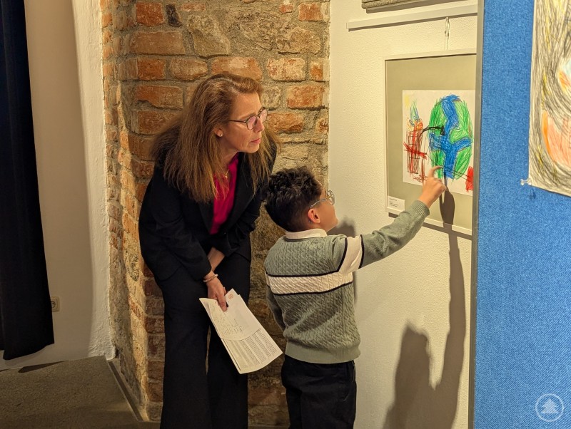 Einer der Preisträger erklärt Frau Wallstabe-Watermann und allen Anwesenden begeistert, was ihn zu seinem Kunstwerk inspirierte. Kind deutet auf Bild und erklärt Frau, was darauf zu sehen ist