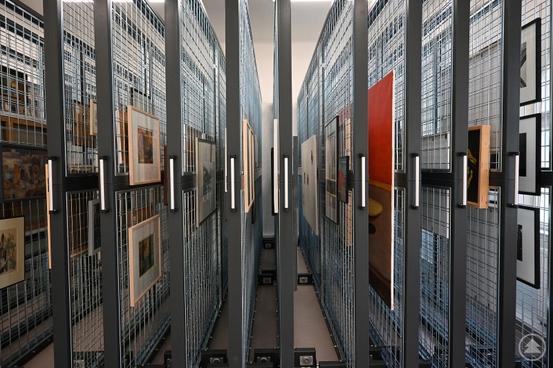 Im Depot der Artothek Niederbayern lagern die Werke fachgerecht an verschiebbaren Gitterwänden. Blick in ein modernes Kunstdepot: Zahlreiche Kunstwerke und gerahmte Bilder hängen ordentlich an verschiebbaren Gitterwänden mit Griffen. Die Werke sind sicher gelagert, das System wirkt geordnet und professionell.
