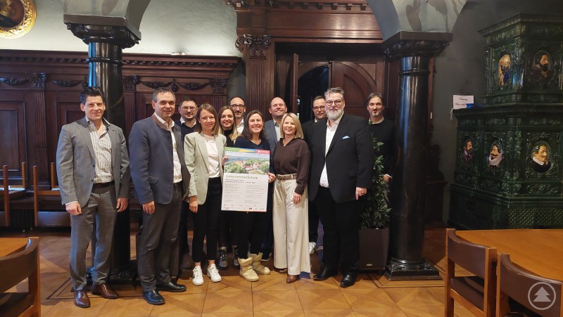 Auftakt zur UnternehmerSchule in Passau: Vertreter der Hans Lindner Stiftung sowie Partner aus Stadt und Landkreis präsentieren die kostenfreie Seminarreihe für Gründer. Gruppenfoto mit mehreren Personen in einem holzvertäfelten Raum. In der Mitte wird ein Plakat zur Seminarreihe UnternehmerSchule in die Kamera gehalten.