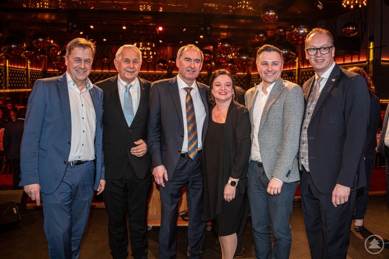 Bürgermeister Alexander Mayer (Grafenau), Kreisrat Herbert Kern, StM Hubert Aiwanger, Nadine Aulinger (Bürgermeisterkandidatin und Gemeinderätin Schöfweg), Marco Braml (Bürgermeisterkandidat und Gemeinderat Gemeinde Saldenburg), MdL Martin Behringer Zwei Männer im Anzug und eine Frau im festlichen Dirndl stehen eng beieinander auf einer Bühne. Im Hintergrund ist ein festlich beleuchteter Saal mit Gästen zu sehen.