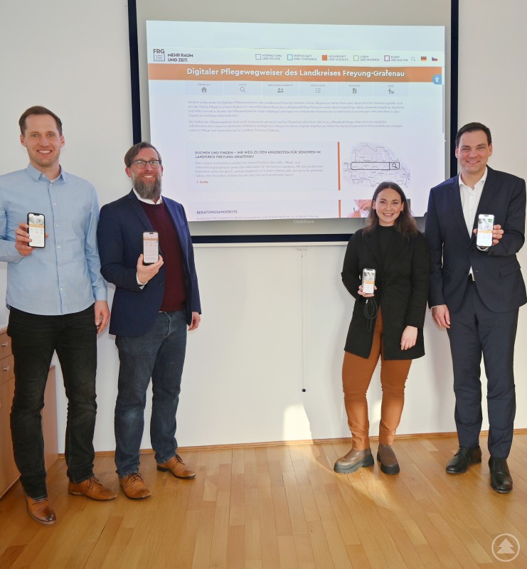 Stellten die neue leicht zugänglichen digitale Plattform zum Thema Pflege und Angebote für Senioren und Menschen mit Handicap im Landkreis vor von rechts vor: Landrat Sebastian Gruber, Lena Schandra, Projektleitung Smartes Land ILE Ilzer Land, Christian Fiebig, Sachgebietsleiter Landkreisangelegenheiten und René Kurtz, Leitung GesundheitsregionPlus Landkreis Freyung-Grafenau. Vier Personen stehen vor einer Präsentationsleinwand und halten Smartphones in der Hand. Auf der Leinwand ist der digitale Pflegewegweiser des Landkreises Freyung-Grafenau zu sehen.