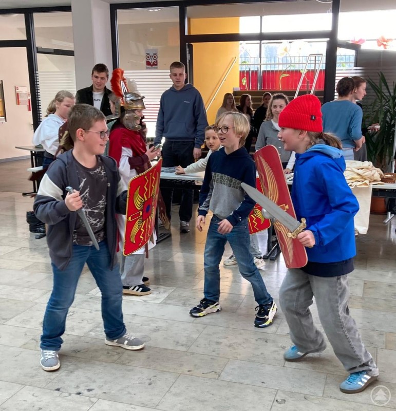 Mehrere Sch&uuml;lerinnen und Sch&uuml;ler probieren mit Schilden und Schwertern spielerisch r&ouml;mische Rollen aus. Im Hintergrund beobachten weitere Jugendliche das Geschehen.