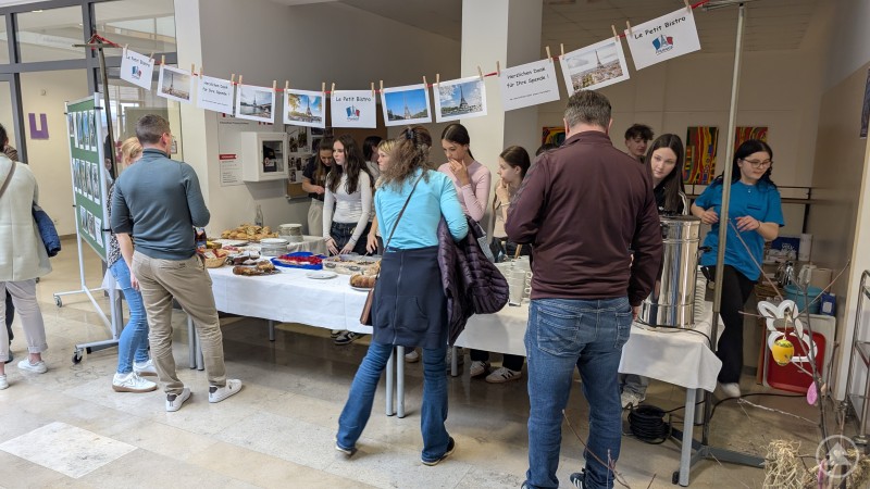 Besucher stehen an einem bewirteten Tisch im Schulgeb&auml;ude, an dem Speisen und Getr&auml;nke ausgegeben werden.