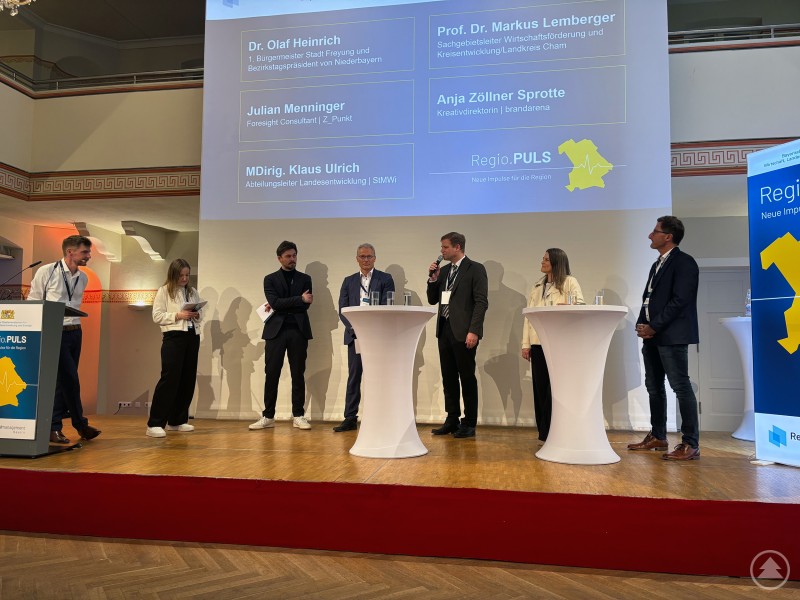 Das Team des Regionalmanagements Freyung-Grafenau mit Regionalmanager Stefan Schuster und der Projektassistenz Chiara Löw präsentierte Staatsminister Aiwanger die Ergebnisse der Arbeitsgruppe „Regionale Identität“. Mehrere Personen stehen auf einer Bühne bei einer Podiumsdiskussion, im Hintergrund ist eine Präsentationsleinwand mit dem Titel der Veranstaltung zu sehen.