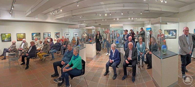 Dicht gedrängt standen und saßen die Gäste bei der Vernissage in St. Oswald. Das zweite Bild zeigt einen voll besetzten Ausstellungsraum mit zahlreichen Besuchern, die größtenteils sitzen und aufmerksam nach vorne blicken – vermutlich auf eine Präsentation oder Rede. Die Ausstellung besteht aus Fotografien an den Wänden sowie gläsernen Vitrinen mit kunstvoll gestalteten Objekten, vermutlich Keramik oder Glas. Die Atmosphäre wirkt feierlich und interessiert, was auf eine Vernissage oder eine besondere Veranstaltung hinweist.