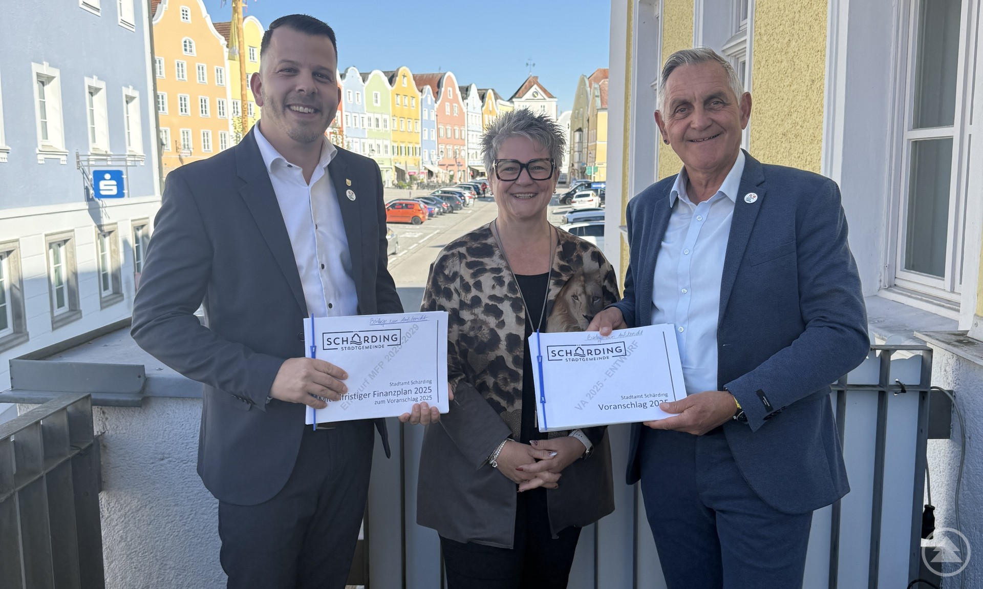 Bürgermeister Günter Streicher, Finanzabteilungsleiterin Martha Quirchtmaier und Finanzstadtrat Paul Königsberger präsentieren den Voranschlag 2025 der Stadt Schärding. Drei Personen stehen auf einem Balkon vor einer bunten Häuserzeile. Zwei Männer im Anzug und eine Frau in gemusterter Jacke halten je eine Mappe mit dem Titel „Stadtgemeinde Schärding Voranschlag 2025“ in den Händen und lächeln in die Kamera.
