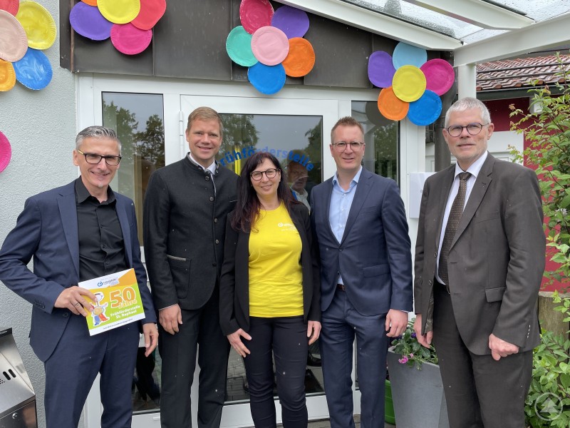Bezirkstagspräsident Dr. Olaf Heinrich (2.v.l.) und der Leiter der Sozialverwaltung des Bezirks, Alexander Seidl (2.v.r.), gratulierten Lebenshilfe-Vorsitzendem Wolfgang Geier (r.), LH-Geschäftsführer Volker Kuppler (l.) sowie der Leiterin der Frühförderstelle, Margit Grill, zum 50-jährigen Jubiläum Fünf Personen stehen vor dem Eingang einer Einrichtung mit bunten Blumen-Dekorationen aus Papptellern. Die mittlere Person trägt ein gelbes Shirt, die anderen Anzug oder Sakko. Eine Person hält ein Schild mit der Aufschrift „50 Jahre Frühförderstelle St. Raphael“.