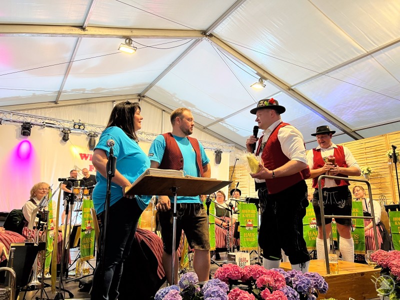 Begrüßung und Grußworte durch die Vorsitzende der Musikkapelle Schnetzenhausen. Mehrere Musikerinnen und Musiker auf einer Bühne im Zelt, zwei Personen in blauen Shirts sprechen in Mikrofone.