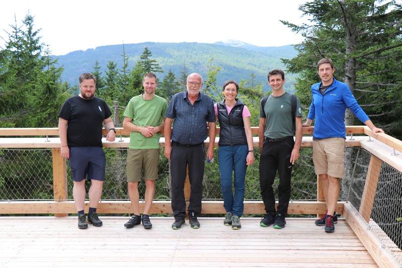 Genießen den Lusenblick an der Aussichtsplattform: Nationalparkleiterin Ursula Schuster mit Bürgermeister Heiner Kilger (v.l.), Till Clos, Leiter der Nationalparkdienststelle Finsterau, Max Greiner, Vorsitzender von Pro Nationalpark, Josef Štemberk vom Nationalpark Šumava sowie Johannes Dick, Bauingenieur des Nationalparks Sechs Erwachsene stehen auf einer hölzernen Aussichtsplattform mit Blick auf bewaldete Berge im Hintergrund.