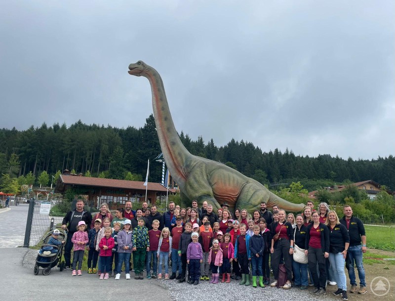 Gemeinsam stark: Die 65 Teilnehmer des Familienausflugs am Eingang des Freizeitparks Edelwies – mit (fast) echtem Dino im Hintergrund. Eine große Gruppe von Erwachsenen und Kindern posiert vor dem Eingang des Freizeitparks Edelwies. Im Hintergrund steht ein riesiger Dinosaurier-Nachbau.