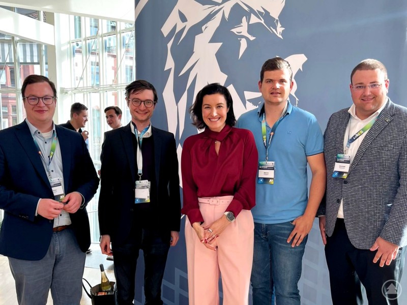 Die Delegation der Jungen Union Freyung-Grafenau bei der 75. Landesversammlung der JU Bayern in Aschaffenburg: Die Delegierten Sebastian Graup (2. v. li.) und Martin Höppler (2. v. re.) sowie Kreisvorsitzender Christoph Weishäupl (1. v. re.) mit dem niederbayerischen Bezirksvorsitzenden Julien Pursch (1. v. li.) und Bundesministerin Dorothee Bär (Mitte). Fünf Personen stehen nebeneinander vor einem blauen Hintergrund mit Löwenmotiv. Sie tragen Namensschilder und lächeln in die Kamera, die mittlere Person trägt ein weinrotes Oberteil.