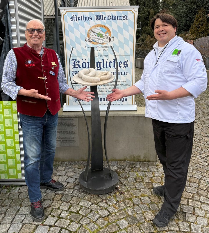 Im Bild (v.l.n.r.): Bertl Fritz, Bayerischer Weißwurstbotschafter und Stefan Einsle, Obermeister der Metzger-Innung ArberLand Zwei Männer präsentieren ein Denkmal mit stilisierten Weißwürsten auf einem Marmorteller. Im Hintergrund ist ein Schild mit dem Schriftzug Mythos Weißwurst und ein Seminarhinweis zu sehen. Beide Männer lächeln und tragen traditionelle bzw. Metzgerkleidung.