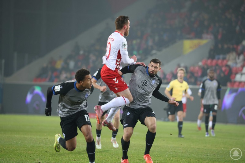 Ein Spieler des SSV Jahn Regensburg steigt im Zweikampf mit zwei Gegenspielern des SV Waldhof Mannheim hoch zum Ball, intensiver Luftkampf im Flutlichtspiel.