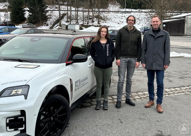 Freuen sich über die Führerscheinförderung für Mitarbeitende ambulanter Pflegedienste (v. l.): Danis Haijnalka, Mitarbeiterin der Ambulanten Pflege Rosenium, Michael Schmid, Regionalleiter der Ambulanten Pflege Rosenium in Niederbayern, und René Kurtz, Leiter der GesundheitsregionPlus Landkreis Freyung-Grafenau. Drei Personen stehen im Freien neben einem Dienstfahrzeug eines ambulanten Pflegedienstes. Sie posieren lächelnd für ein Foto auf einem Parkplatz, im Hintergrund sind winterliche Bäume und Gebäude zu sehen.