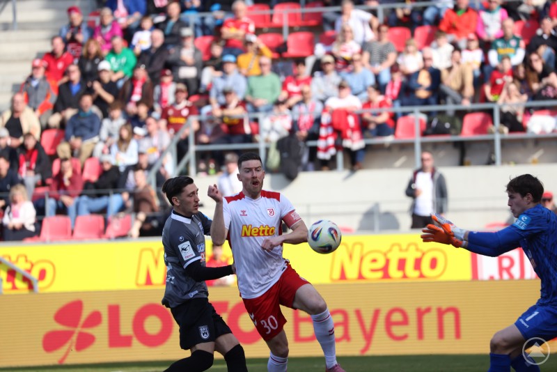 Ein Jahn Spieler nimmt den Ball im Strafraum an, w&auml;hrend ein Ulmer Gegenspieler und der Torh&uuml;ter eingreifen wollen.