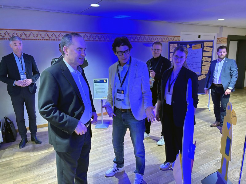 Bezirkstagspräsident Dr. Olaf Heinrich appellierte im Rahmen einer Podiumsdiskussion an Akteurinnen und Akteure der Regionalentwicklung, mutig voranzugehen. Eine Gruppe von Teilnehmern steht in einem Ausstellungsbereich und betrachtet gemeinsam eine Präsentation, während sie sich austauschen.