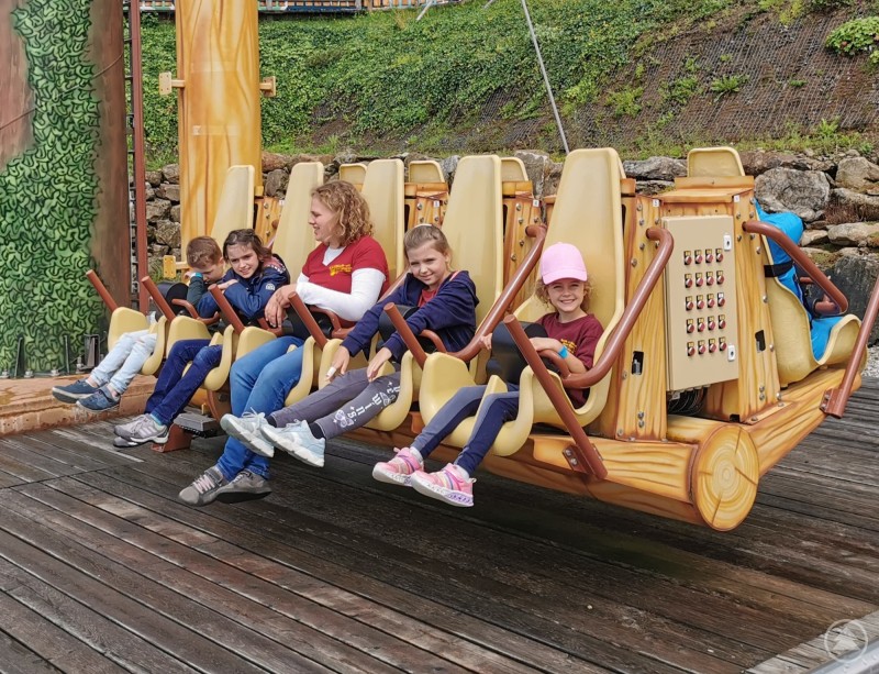 Kinder sitzen nebeneinander in einer Holzschaukel-Attraktion und werden seitlich geschaukelt.